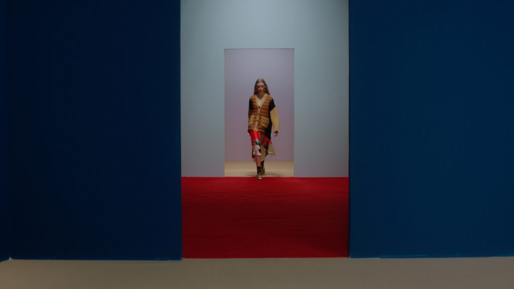Color grading example from a profesional film colorist. Young blonde woman dressed in brown and red, walking in a hallway, blue, light blue and lilac, with a red floor.